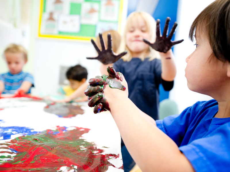 Little boy doing messy play with paint. A little girl showing her paint stained hands together with 2 little boys can be seen in blurred background.