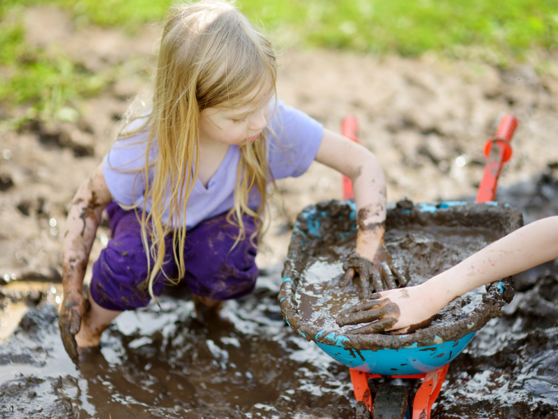A little girl enjoying messy play with mud, placing mud in a toy wheel borrow. 